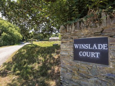 A stone wall with a Winslade Court sign next to a country road with trees and a building in the background at Cowslip Cottage in Frogmore