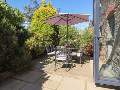 An outdoor patio with a table umbrella and six chairs surrounded by plants and a stone wall at Cowslip Cottage in Frogmore