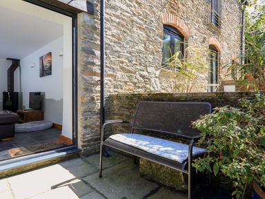 An outdoor patio with a metal bench and cushion next to a stone wall and an open door leading to a living room at Cowslip Cottage in Frogmore