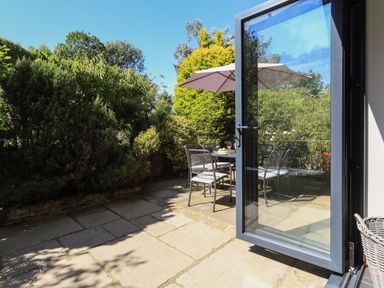 A patio with a table, chairs, and an umbrella next to a garden visible through an open glass door at Cowslip Cottage in Frogmore