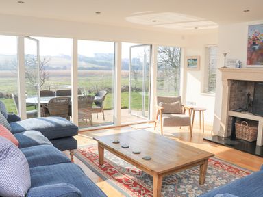A living room with a wooden coffee table blue sofas a rug and a beige armchair next to a fireplace with glass doors opening to a garden at Sea Peep in Thurlestone