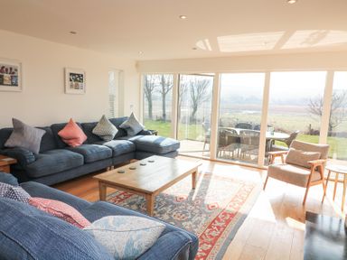 A living room with blue sofas cushions a wooden coffee table and armchair next to large glass doors at Sea Peep in Thurlestone