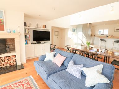 A living room with a blue sofa and pillows a wooden dining table with chairs and a kitchen area at Sea Peep in Thurlestone