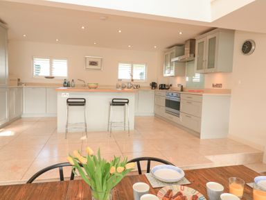 A kitchen with an island and two stools a dining table with cups and plates and yellow flowers in a vase at Sea Peep in Thurlestone