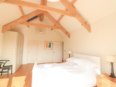 A bedroom with a white bed two bedside tables with lamps a wardrobe a black chair wooden beams on the ceiling and a framed picture on the wall at Sea Peep in Thurlestone