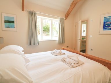 A bedroom with a wooden bed frame white bedding folded towels on the bed a window with curtains and framed artwork on the walls at Sea Peep in Thurlestone