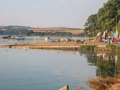 A riverside with boats docked near the shore and kayaks stacked near trees at Sea Peep in Thurlestone