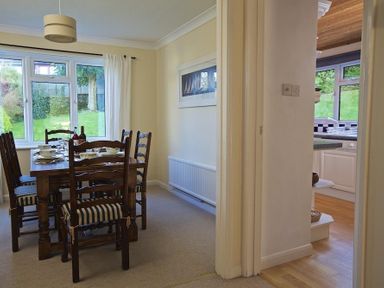 A dining room with a wooden table and chairs set near a window showing a garden and a view of a kitchen area at Amberley in Dartmouth