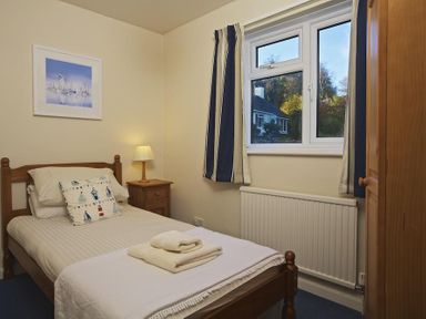 A bedroom with a single bed bedside table with a lamp wardrobe and window with blue and white curtains at Amberley in Dartmouth