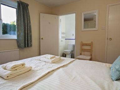 A bedroom with a made bed and towels a window with curtains a door leading to a bathroom with a toilet a wooden chair and a wall mirror at Amberley in Dartmouth