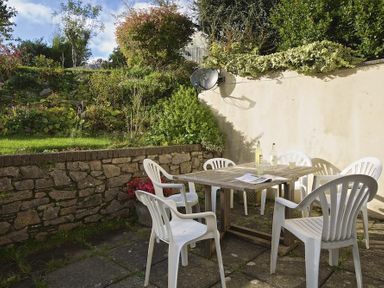An outdoor patio area with a wooden table and six white plastic chairs by a stone wall with plants at Amberley in Dartmouth