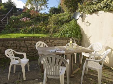 An outdoor patio with a wooden table and six white plastic chairs surrounded by a stone wall and garden at Amberley in Dartmouth
