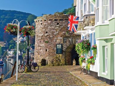 A cobblestone walkway along water with hanging flower baskets a bicycle a stone archway and buildings with flowers at Amberley in Dartmouth