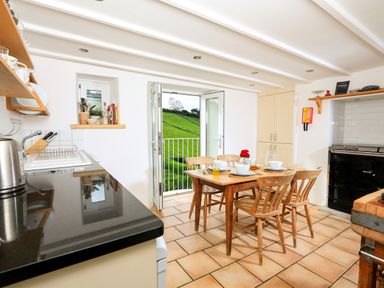 A kitchen with a wooden dining table and chairs set with dishes and glasses and open doors revealing a green grassy hill outside at Bow Cottage in East Portlemouth