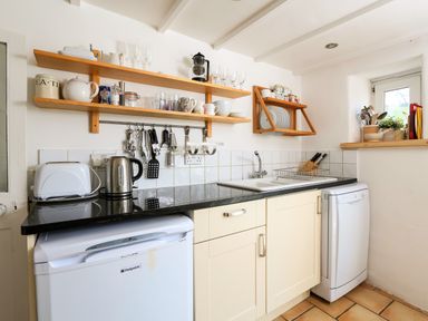 A kitchen with shelves holding cups and glasses a toaster kettle utensils and a sink with a drying rack at Bow Cottage in East Portlemouth