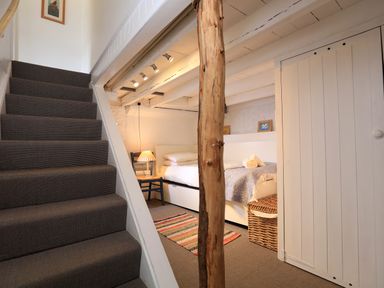 A small bedroom with a single bed a wooden chair with a lamp a striped rug and a wicker basket under stairs at Bow Cottage in East Portlemouth