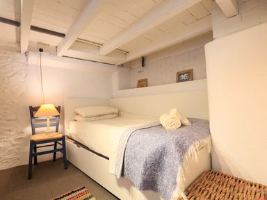 A small bedroom with a single bed a blue blanket two rolled towels a wooden chair with a lamp and framed pictures on a shelf at Bow Cottage in East Portlemouth