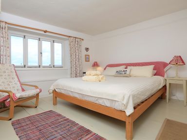 A bedroom with a wooden bed with white bedding and rolled towels a chair with floral cushions and a window with curtains at Bow Cottage in East Portlemouth
