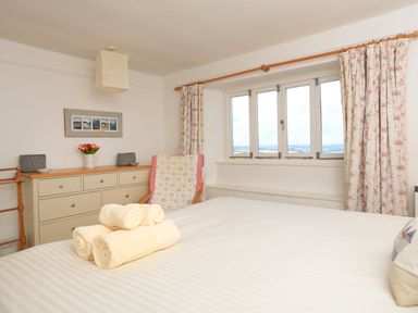 A bedroom with a bed, rolled towels, dresser with flowers, chair, window with curtains at Bow Cottage in East Portlemouth