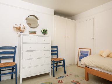 A bedroom with a white dresser between two blue chairs a bed with beige bedding and a framed picture on the floor at Bow Cottage in East Portlemouth