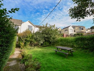 A garden with a wooden picnic table a grassy lawn and a pathway beside bushes and houses in the background at Bow Cottage in East Portlemouth
