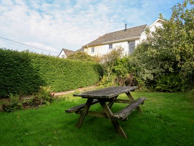A garden with a wooden picnic table surrounded by bushes and a house in the background at Bow Cottage in East Portlemouth