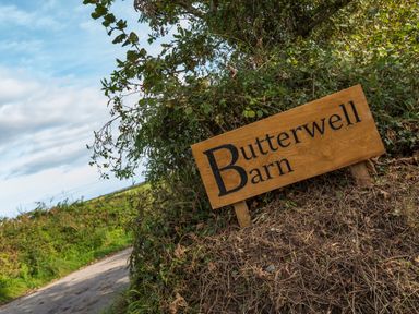 A wooden sign with Butterwell Barn written on it near a country road surrounded by bushes at Butterwell Barn in Dartmouth