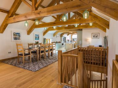 A dining area with a wooden table and chairs under exposed wooden beams next to a kitchen with mint green cabinets at Butterwell Barn in Dartmouth