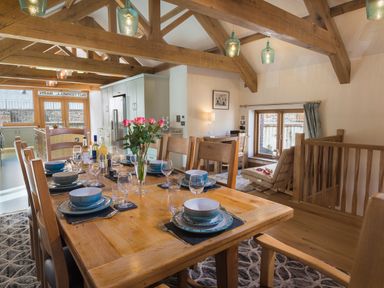 A dining area with a wooden table set with plates bowls glasses and cutlery a kitchen in the background and a seating area with a chair and window at Butterwell Barn Dartmouth