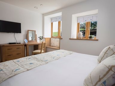 A bedroom with a bed facing a dresser and mirror beside two windows with patterned blinds at Butterwell Barn in Dartmouth