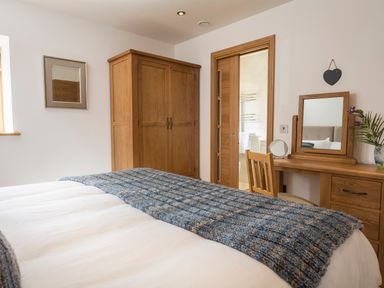 A bedroom with a bed covered in white sheets and a blue knitted throw blanket a wooden wardrobe a wooden dressing table with mirror and chair and a door to an en suite bathroom at Butterwell Barn in Dartmouth