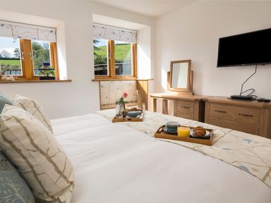 A bedroom with a bed breakfast trays wooden dresser mirror wall mounted tv and windows with a view at Butterwell Barn in Dartmouth