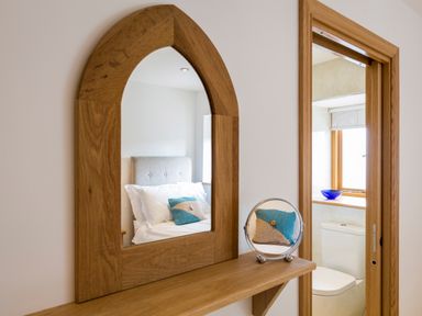 A mirror with wooden frame reflecting a bed with pillows and a bathroom with a toilet and window at Butterwell Barn in Dartmouth