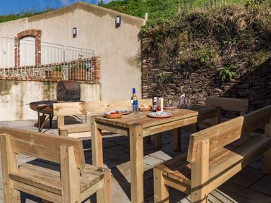 An outdoor patio with a wooden table and benches set with plates bottles and glasses near a barbecue grill at Butterwell Barn in Dartmouth
