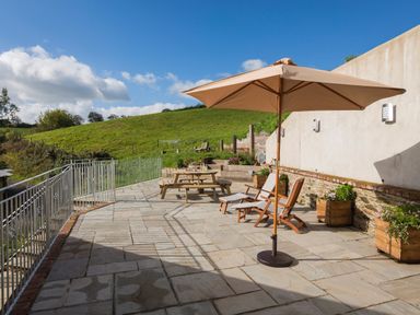 An outdoor patio with a large umbrella wooden chairs and a picnic table overlooking a grassy hill at Butterwell Barn in Dartmouth