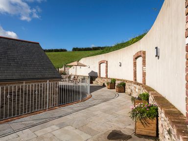 An outdoor terrace with stone flooring wooden flower boxes a curved wall and a roofed structure at Butterwell Barn in Dartmouth