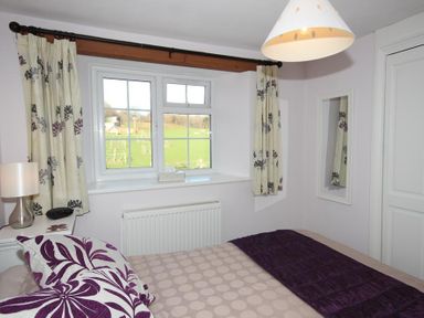 A bedroom with a bed a window with curtains a bedside table with a lamp and a mirror on the wall at Cloverwell in Woodleigh