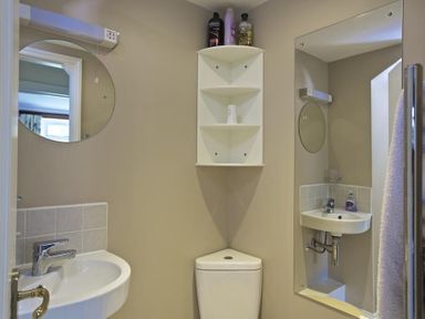 A bathroom with a corner toilet a wall-mounted sink round mirror and a white corner shelf holding toiletries at Cloverwell in Woodleigh