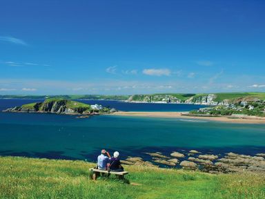 A coastal scene with two people sitting on a bench overlooking the ocean and cliffs at Cloverwell in Woodleigh