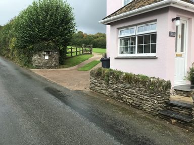 A pink cottage with a white door and window next to a stone wall and a green hedge by a narrow road at Cloverwell in Woodleigh