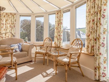 A sunroom with wicker chairs a small glass table a beige armchair and floral curtains at Cottage View in Hope Cove