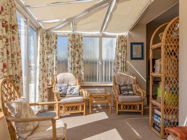A sunroom with wicker chairs pillows a wicker bookshelf and floral curtains at Cottage View in Hope Cove