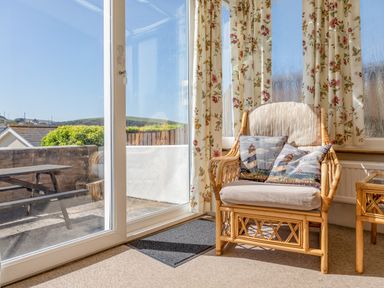 A room with a wicker armchair and floral curtains next to a glass sliding door and a wooden picnic table outside at Cottage View in Hope Cove