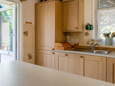 A kitchen with wooden cabinets a sink a blender a bread box utensils in a container and an open door at Cottage View in Hope Cove