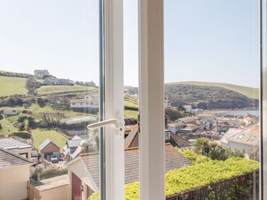 A view through a closed window showing houses, green hills, a small town, and the sea at Cottage View  Hope Cove