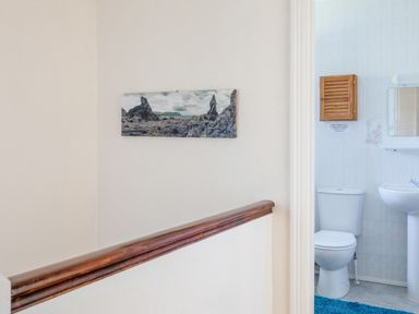 A hallway with a wooden handrail and a wall photo of rocks leading to a bathroom with a white toilet sink and a wooden cabinet at Cottage View in Hope Cove