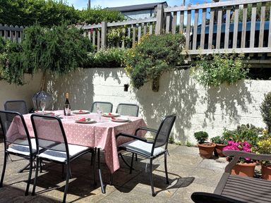 A patio dining area with a table covered by a pink polka dot tablecloth set with plates and glasses and surrounded by metal chairs near a white wall with plants at Cottage View in Hope Cove