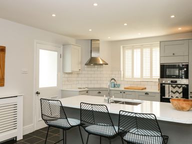 A kitchen with three black wire bar stools at an island with a sink and white countertop at Cindy's Retreat in Hope Cove