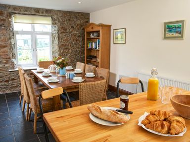 A dining room with a wooden table set for eight with bowls and mugs a kitchen counter with bread croissants and orange juice at Cindys Retreat in Hope Cove