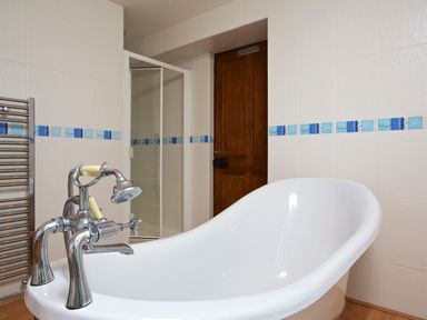 A bathroom with a white bathtub with chrome faucet a wooden door a glass shower enclosure and blue tile accents on the walls at Cindys Retreat in Hope Cove